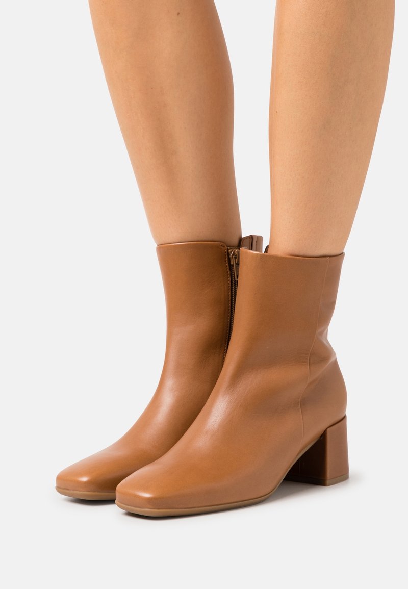 Bottes cheville en cuir fauve avec talons carrés portées sur jambes nues, dotées de fermetures éclair latérales et d'une finition lisse et polie.