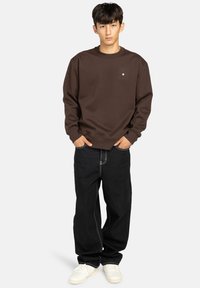 Felpa marrone in cotone con scollatura rotonda e un piccolo logo sul petto, abbinata a jeans neri a gamba larga e sneakers bianche.