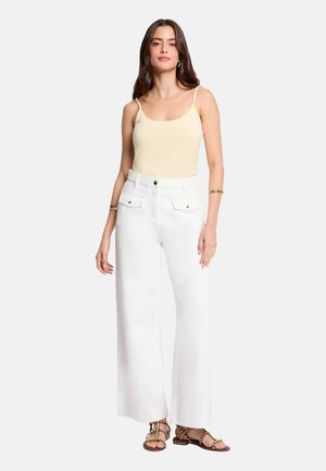 Femme portant un débardeur crème, un pantalon blanc à jambes larges et taille haute, et des sandales marron cloutées, debout devant un fond uni.