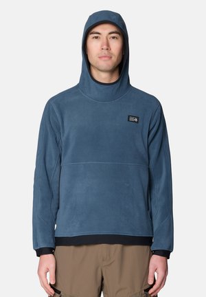 Mann trägt einen blauen Kapuzen-Fleece-Sweatshirt und braune Hose, steht frontal vor einem einfarbigen Hintergrund.
