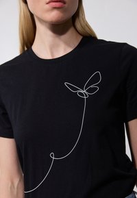 Svart t-skjorte laget av bomull med rund hals, med et hvitt linjært sommerfugl-design på venstre side. Glatt tekstur.