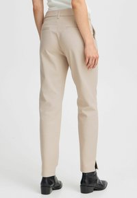 Pantalons beige ajustés à coupe droite, dotés de deux poches arrière, de fentes sur le bas et portés avec des bottines noires à talons.