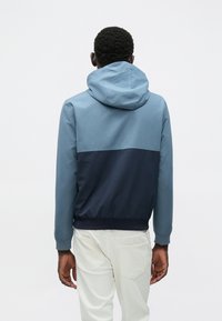 Μπουφάν με κουκούλα με σχεδιασμό σε χρωματικούς τομείς, με ανοιχτό μπλε ανώτερο τμήμα και σκούρο ναυτικό μπλε κατώτερο τμήμα. Το ύφασμα φαίνεται λείο και ματ.