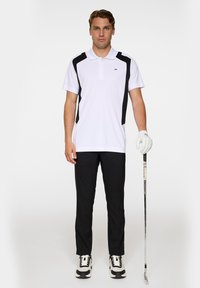 Vit polo med svarta sidopaneler, kortärmad och med krage, kombinerad med svarta byxor och svart-vita golfskor, håller en golfklubba.