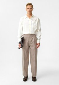 Witte overhemd met lange mouwen en knoopsluiting, beige plooibroek, bruine schoenen, houdend een donkere clutch, staande op een effen achtergrond.