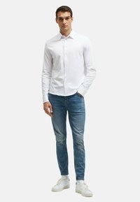 Weißes Hemd mit Knöpfen, blaue Jeans und weiße Sneaker. Moderner Schnitt mit einer glatten Baumwolltextur. Neutrale Kulisse hebt das Outfit hervor.
