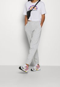 Przycięty biały t-shirt z kolorową grafiką tęczy, jasnoszare spodnie dresowe z kieszeniami oraz kolorowe sneakersy New Balance z odblaskowymi elementami.