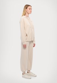 Beige mikina s kapucí, zipem, stahovací šňůrkou a bočními kapsami, v kombinaci s odpovídajícími bežovými joggerami s elastickými manžetami a minimalistickým designem.