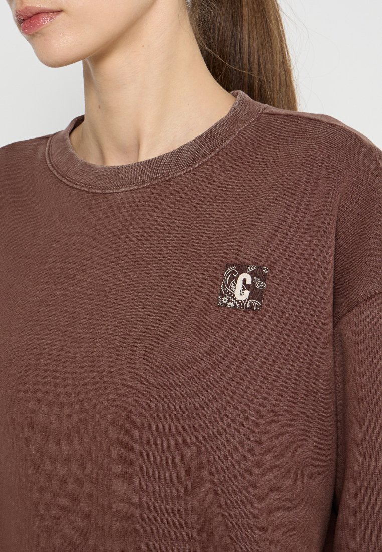 Brauner Sweatshirt mit rundem Ausschnitt und gerippten Bündchen. Auf der Vorderseite befindet sich ein kleiner quadratischer Patch mit Paisley-Design.