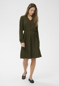 Olivno zelena obleka z V-izrezom, dolgimi rokavi in pasom za zavezovanje v pasu. Mehka, teksturirana tkanina; krilo sega do nekoliko nad kolena. Oblečena s črnimi balerinkami.