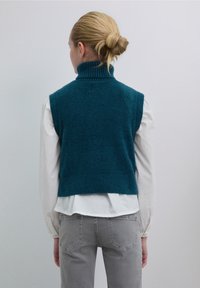 Grünblauer ärmelloser Pullover mit hohem Kragen, aus weichem Material, mit kurzem Schnitt und glatter Textur, über einem weißen Hemd getragen.