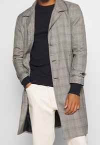 Grå rutig kappa med slagkrage, som har två framfickor, knäppta ärmar och en mjuk textur, lagd över en mörk ribbad tröja.