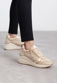 Las zapatillas de plataforma beige presentan una textura acolchada, acentos metálicos, una cremallera decorativa en el lateral y un diseño de cordones. Combinadas con leggings negros.