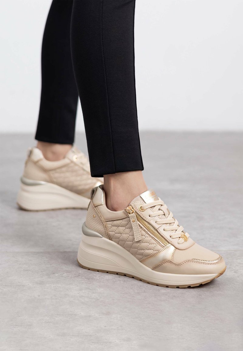 Las zapatillas de plataforma beige presentan una textura acolchada, acentos metálicos, una cremallera decorativa en el lateral y un diseño de cordones. Combinadas con leggings negros.