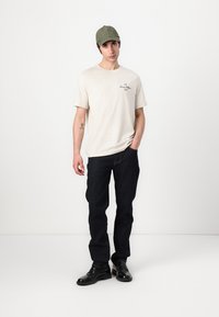 Beige short-sleeve t-shirt with printed text, dark blue jeans, black shoes, and a green cap. Flat footwear, minimalistic design.
