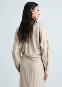 Beige knitted top with long sleeves, relaxed fit, soft texture, and a rounded hem. Worn with a matching bottom. Gold hoop earrings visible.
