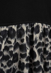 Schwarzes Oberteil mit einem gemusterten Beigen und schwarzen Leopardenmuster-Rüsche. Der Stoff hat eine unterschiedliche Textur zwischen glatt und strukturiert. Sichtbare Nahtdetails.