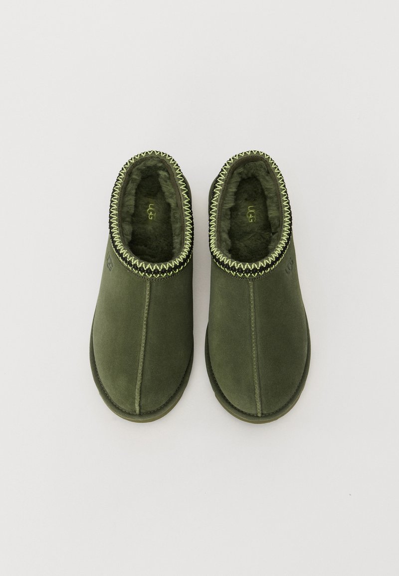 Groene suède instappers met schapenvachtvoering, contrasterende zigzagsteek langs de bovenrand en een platte rubberen zool.