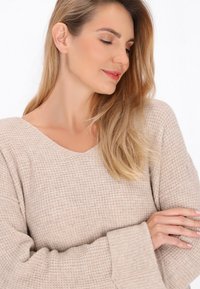 Maglione beige lavorato a maglia con scollatura ampia e maniche lunghe leggermente svasate. Texture morbida e dettaglio di motivo sottile sul tessuto.