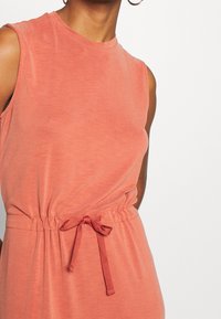 Persikka värinen hihaton mekko, jossa on pyöreä kaula-aukko, kapea vyötärö ja nauhoilla sidottu yksityiskohta, joka on valmistettu pehmeästä, teksturoidusta kankaasta.