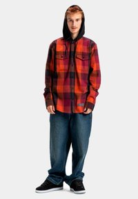 Camisa de franela a cuadros rojos y negros con capucha negra, que presenta dos bolsillos en el pecho y un diseño de botones, combinada con jeans azules holgados y zapatillas negras.