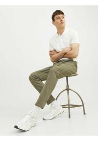 Witte polo, olijfgroene broek, witte sneakers, zittend op een metalen kruk. Eenvoudige achtergrond, ontspannen houding, gladde stoftextuur.