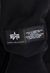 Zwarte stoffen tag met het Alpha Industries-logo en witte tekst met de vermelding Knoxville, Tennessee, VS, opgericht oktober 1959, details van de aannemer van het Amerikaanse Ministerie van Defensie.