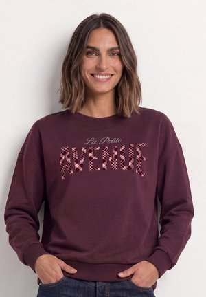 Kvinde med skulderlangt brunt hår, smilende, iført en mørkerød sweatshirt med teksten "La Petite Avenue" og hænderne i lommerne.