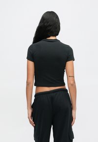 Γυναίκα με μακριά μαύρα μαλλιά, φορώντας κοντό μαύρο μπλουζάκι και μαύρο παντελόνι cargo, στέκεται γυρισμένη με την πλάτη σε έναν απλό φόντο.