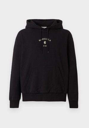 Man standing wearing black hoodie with front pocket and “MARGIELA 6” logo, wide black pants, and black shoes against plain background.