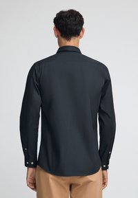 Maglietta nera a maniche lunghe con colletto appuntito, texture liscia e polsini con bottoni. Presenta un design posteriore pulito e sartoriale.