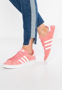 Persona che indossa sneakers Adidas Campus rosa e jeans blu con una striscia in denim chiaro lungo il lato, in piedi su un pavimento bianco.