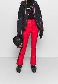 Zwarte ski-jas met rode accenten, nauwsluitende rode skibroek met zwarte zijkstrepen en stiksels, zwarte helm en skischoenen.