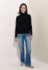 Svart stickad tröja med hög hals och snöre, ihop med ljusblå utsvängda jeans och beige skor. Slät textur och kroppsnära design.