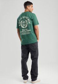 T-shirt verde in cotone con stampa grafica bianca sul retro che presenta un design di un globo e un lauro. Abbinato a jeans grigi scuri e scarpe bianche.