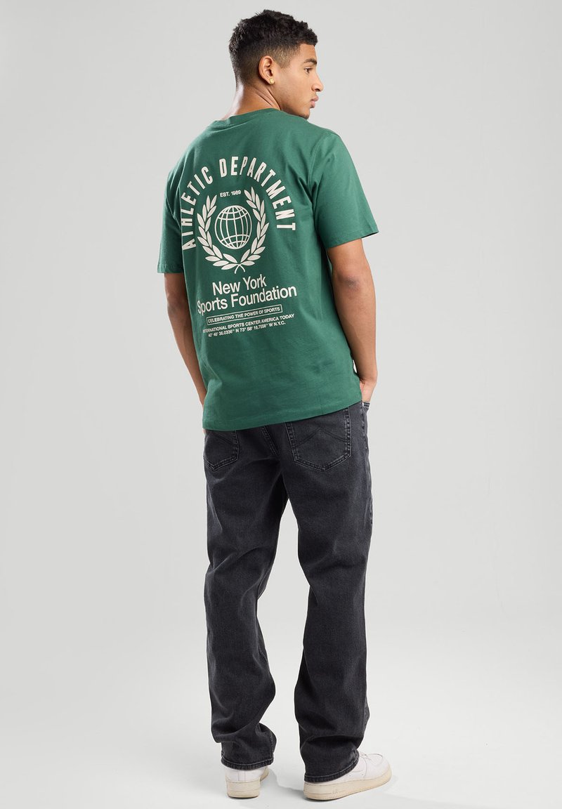 T-shirt verde in cotone con stampa grafica bianca sul retro che presenta un design di un globo e un lauro. Abbinato a jeans grigi scuri e scarpe bianche.