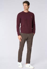 Maglione a treccia bordeaux con polsini a coste, abbinato a chinos marroni e sneaker bianche, in piedi su uno sfondo neutro.