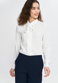 Biała bluzka z długim rękawem z wiązanym detalem przy szyi, w połączeniu z ciemnonawojskowymi spodniami. Gładki materiał z zapięciem na guzik przy mankietach.