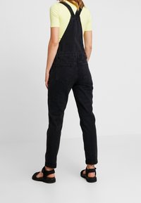 Svart denimoverall med justerbara axelremmar, två bakfickor och avsmalnande ben. Matchat med en ljusgul ribbad tröja och svarta sandaler.