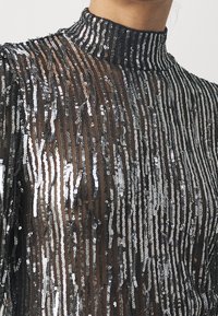 Schwarzes durchsichtiges Oberteil mit vertikalen silbernen Pailletten, hohem Kragen und strukturierter Oberfläche, das ein Muster aus Streifen und dezentem Schimmer hervorhebt.