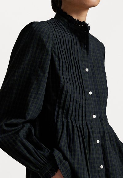 Ciemnozielona koszula w kratę z pionowymi fałdami, bufiastymi rękawami, wysokim koronkowym kołnierzykiem i białymi guzikami wzdłuż zapięcia. Teksturowany materiał.