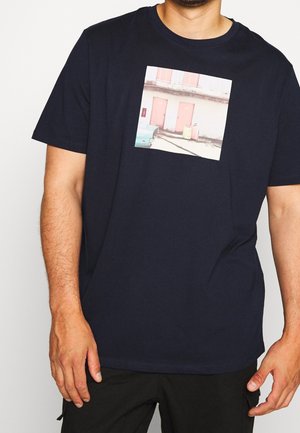 Homme portant un t-shirt bleu marine avec une photo imprimée représentant deux portes roses, des bagages et une voiture vintage sur le devant.