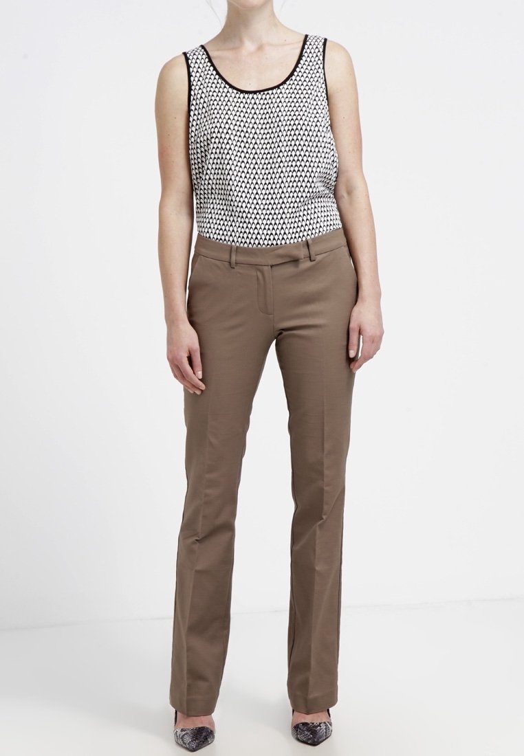 Femme portant un haut sans manches à motif noir et blanc, un pantalon marron à jambes droites, et des chaussures pointues au motif peau de serpent, debout devant un fond blanc.