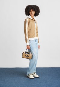 Καφέ μπουφάν με φερμουάρ και λευκές ρίγες, χαλαρά ανοιχτό μπλε τζιν και λευκά αθλητικά παπούτσια. Μικρή καστανομπεζ τσάντα χειρός με μαύρη λεπτομέρεια λογότυπου.