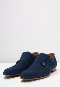 Chaussures en daim bleu marine avec doubles boucles, bout en amande et intérieur en cuir lisse. Semelle en bois plate avec talon minimal.