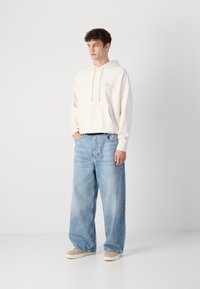 Hoodie beige con coulisse e piccolo logo, abbinato a jeans leggeri blu chiaro. Il modello è in piedi su uno sfondo neutro, indossando scarpe beige.
