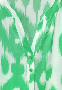 Grüne Bluse mit V-Ausschnitt, die ein Leopardenmuster in verschiedenen Grüntönen zeigt. Enthält sechs passende grüne Knöpfe an der Vorderseite.