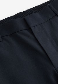 Pantalon noir à taille élastique avec passants de ceinture et fermeture devant en tissu, montrant un gros plan des coutures et de la texture du tissu.