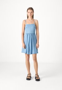 Robe en denim à fines bretelles, corsage ajusté et jupe évasée en bleu clair. Assortie avec des sandales à plateforme noires et des accessoires minimalistes.