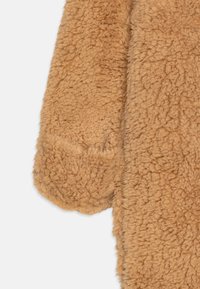 Miękka tkanina w jasnobrązowym kolorze o puszystej fakturze. Posiada zaokrąglony mankiet i lekko luźny fason, odpowiedni na wierzchnią odzież.